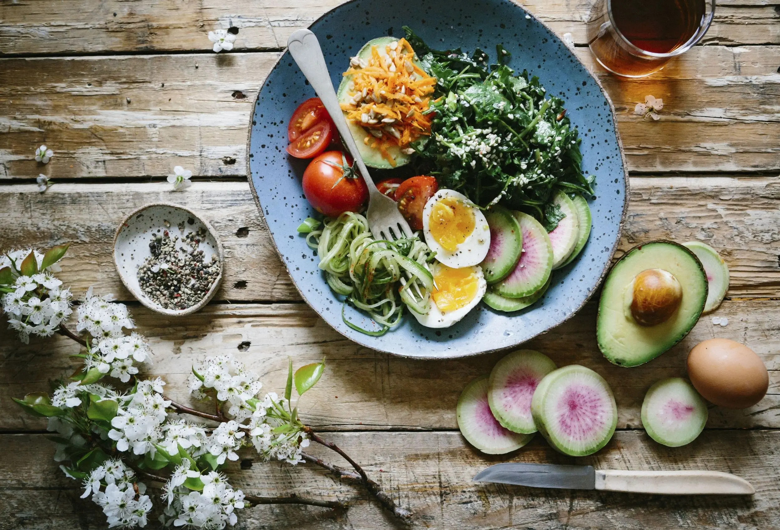 An overhead view of a vibrant bowl of food with eggs, greens, tomatoes, and radishes on a wooden table.